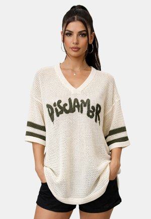 Donna con capelli lunghi e scuri che indossa un maglione bianco a V in maglia con il testo "DISCLAIMER" e pantaloncini neri, in piedi con le mani nelle tasche.