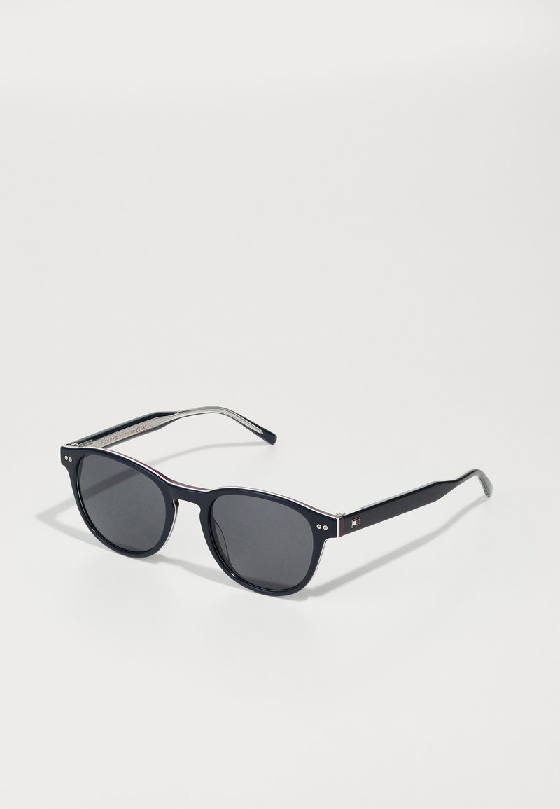 Óculos de sol redondos com lentes escuras, armação de plástico preta, detalhes em metal prateado e uma fina linha vermelha na borda superior.