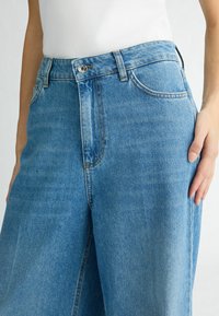 Jeans in denim azzurro chiaro con vita alta, tasche davanti e una vestibilità morbida. Presentano cuciture classiche e una chiusura con bottone in metallo.