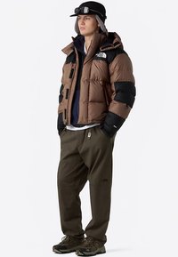 Braune und schwarze Steppjacke mit Reißverschluss vorne, mehreren Taschen und Kapuze. Olivgrüne Hose und dunkle Schuhe vervollständigen das Outfit.