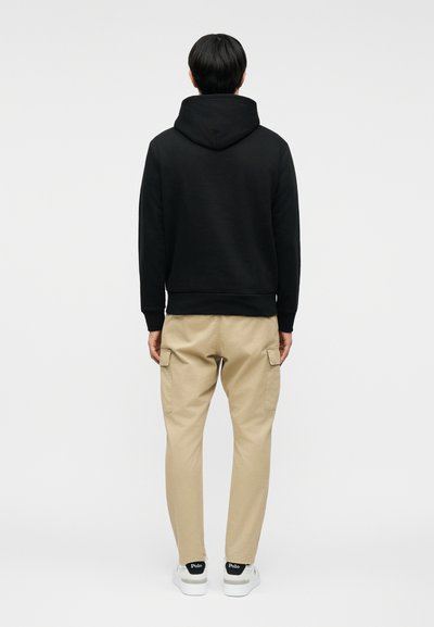 Άτομο που στέκεται με την πλάτη, φοράει μαύρο φούτερ με κουκούλα, μπεζ παντελόνι cargo και λευκά αθλητικά παπούτσια με μαύρη ένδειξη "Polo".