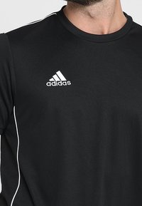 Svart atletisk tröja i texturerad tyg, med rund halsringning och vitt Adidas-logotyp på övre vänstra bröstet. Subtil vit randdekor.