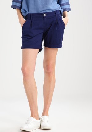 Person iført marineblå shorts med opkrogede kanter, lyseblå top med blondestrimlede ærmer og hvide slip-on sneakers, hænderne i lommerne.