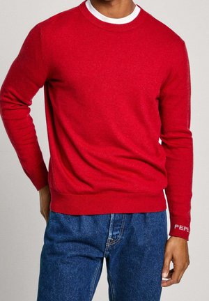 Mann trägt einen leuchtend roten Langarmpullover mit weißem Kragen-Detail und blauen Jeans, Hand auf der Hüfte, gezeigt vom Hals bis zur Hüfte.