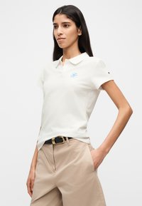 Λευκό μπλουζάκι πόλο με κοντά μανίκια, που διαθέτει ένα ανοιχτό μπλε κεντημένο λογότυπο στο αριστερό στήθος και μια μικρή λεπτομέρεια λογότυπου στο μανίκι.