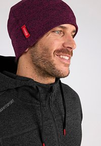 Gorro de punto en magenta oscuro con un acabado texturizado. Presenta una etiqueta roja de marca y se lleva con una sudadera gris con cremallera.