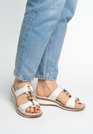 Voeten in witte lederen sandalen met decoratieve kralen, gecombineerd met lichtblauwe jeans die boven de enkels zijn afgeknipt, staand op een witte ondergrond.