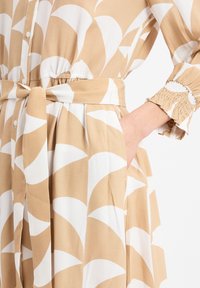 Beige en witte jurk met een patroon, met een strik in de taille, knoopsluiting aan de voorkant, elastische mouwen en zakken; de stof lijkt zacht en lichtgewicht te zijn.