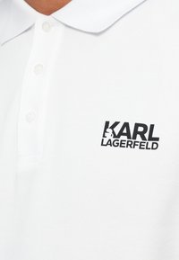 Baltas polo marškinėlis su juodu "KARL LAGERFELD" logotipu ir siluetu kairiajame krūtinės srityje, su apykakle ir trimis mygtukais.