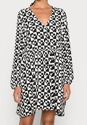 Femme portant une robe à carreaux noir et blanc avec des formes abstraites de cœur, manches longues, col en V et boutons à l'avant.