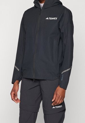 Muž v černé nepromokavé bundě a kalhotách Adidas Terrex stojí s uvolněnými pažemi podél těla před jednobarevným pozadím.