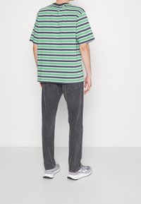 Maglietta oversize a righe verdi, blu e bianche, a maniche corte, abbinata a jeans grigio scuro e sneakers grigie, vista da dietro.