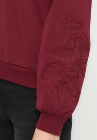 Burgundy sweatshirtärm med upphöjd floral och paisleybroderi, ribbat mudd, buren med svarta byxor mot en vit bakgrund.