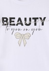 Maglietta bianca con testo in nero e argento in grassetto con la frase "Bellezza Unica." Include un'accenno a forma di fiocco di perline bianche sotto il testo.