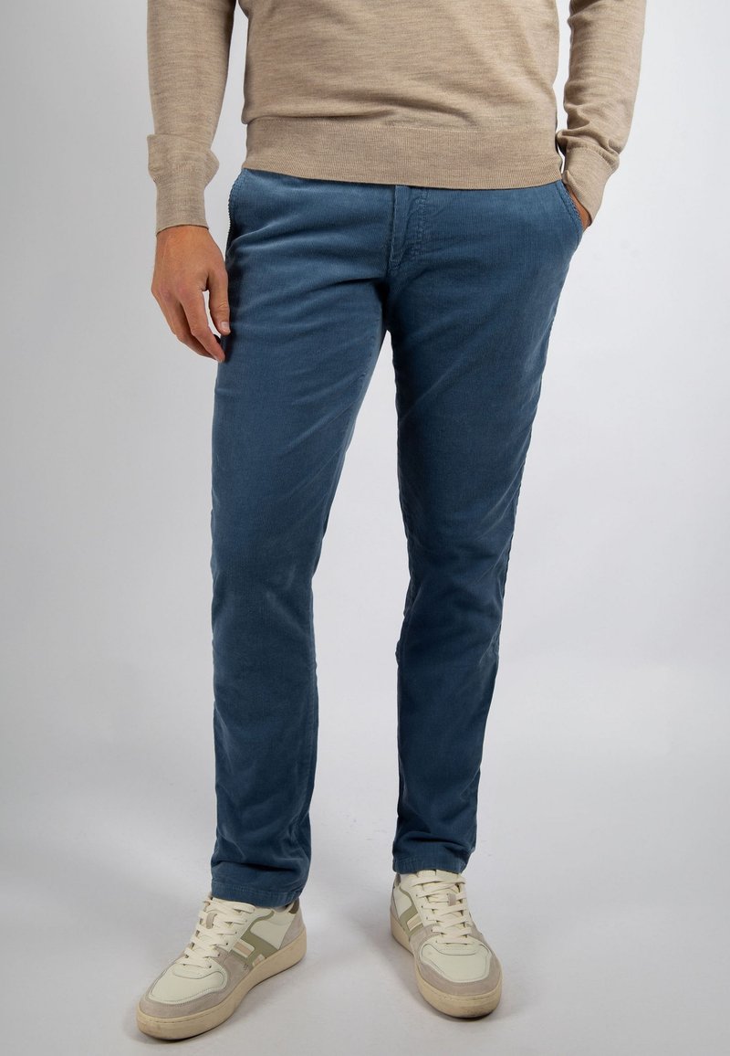 Man met blauwe ribfluwelen broek, beige trui met lange mouwen en witte sneakers, staand met één hand in de zak tegen een effen achtergrond.