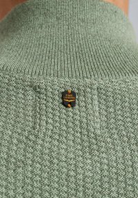 Gebreide groene trui met een textuurpatroon en een klein zwart label met gele tekst "PME LEGEND" bevestigd aan de kraag.