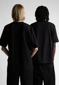 Divi indivīdi stāv viens pret otru, tērpti brīvās formas melnās kokvilnas T-kreklos. Krekliem ir īsas piedurknes un apaļa kakla izgriezuma forma.