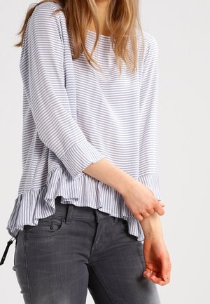 Femme portant un haut à manches longues rayé gris et blanc avec un ourlet à volants et un jean gris, posant les mains près de la taille.