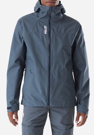 Man wearing a blue waterproof Gore-Tex jacket with a hood, zipped up, and matching pants against a plain background.