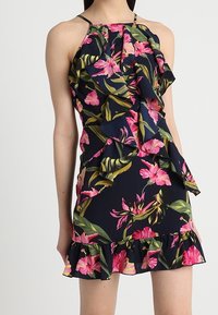 Bezramenné květinové šaty s námořnickým pozadím, velkými růžovými květy a zelenými listy. Obsahují volánové detaily a zprohýbaný lem.