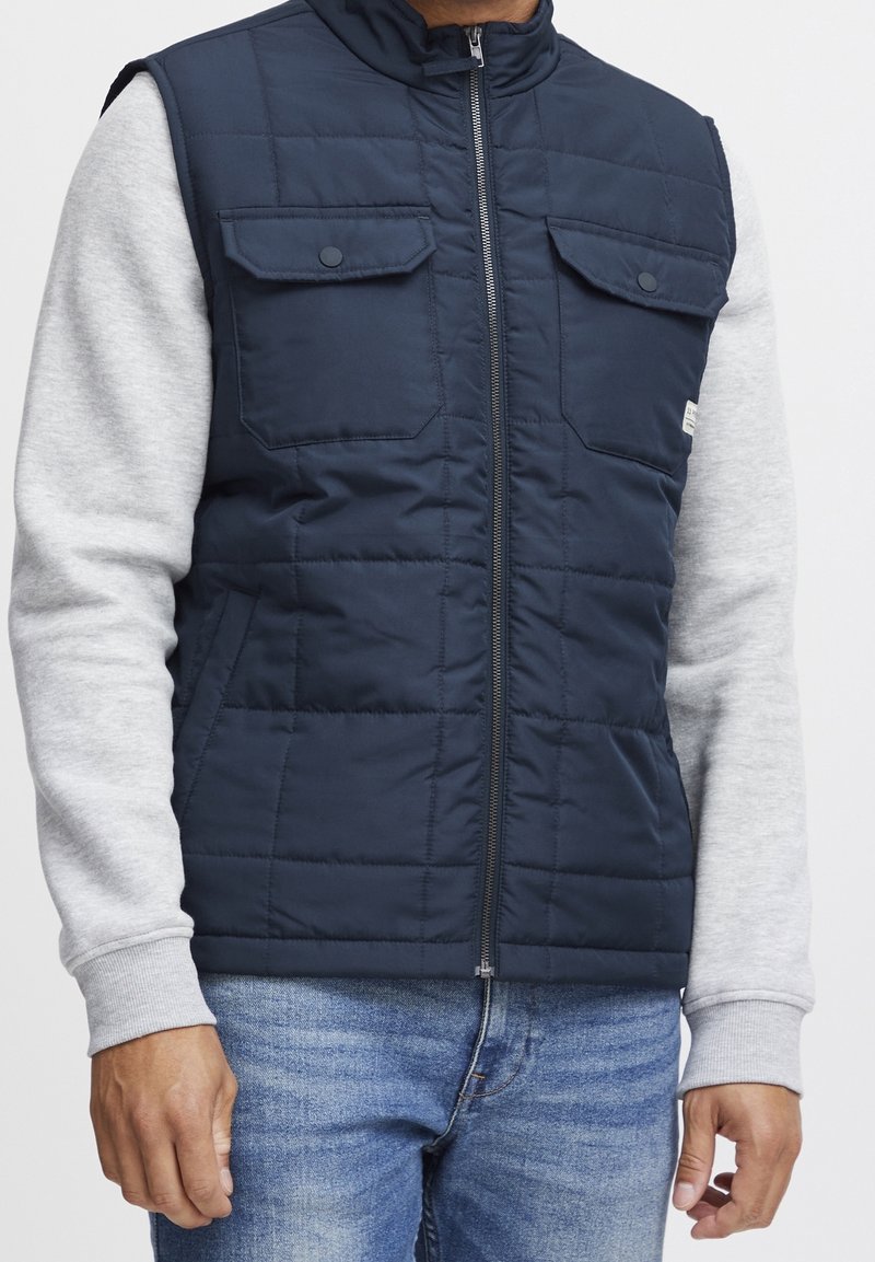 Gilet matelassé bleu marine avec un col montant, deux poches poitrine et une fermeture éclair frontale, porté avec une chemise grise à manches longues. Jean en denim visible.