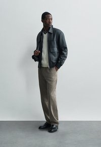 Giacca bomber in pelle nera, maglione testurizzato crema, pantaloni a quadri marroni e scarpe nere, con una silhouette liscia e strutturata.