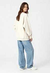 Sweatshirt oversize de couleur crème avec manches longues et poignets côtelés, associé à un jean en denim bleu clair ample et des baskets blanches.