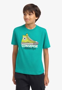 Τυρκουάζ βαμβακερό μπλουζάκι με γραφικό κίτρινο αθλητικό παπούτσι ψηλού τύπου και κείμενο "CONVERSE Athletic Depts." σε λευκό και μαύρο.