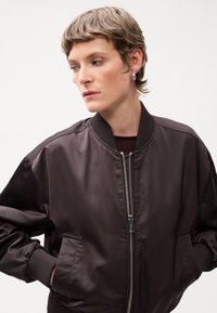 Brown bomber jacket with a zip front, ribbed cuffs, and side pockets. Smooth, shiny material and relaxed fit. Close-up view of wearer.