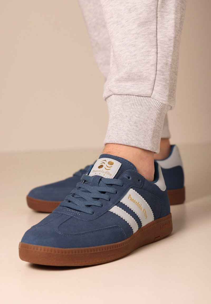 Blaue Wildledersneaker mit weißen Streifen und goldener Markierung, ausgestattet mit einer braunen Gummisohle und Schnürdesign.