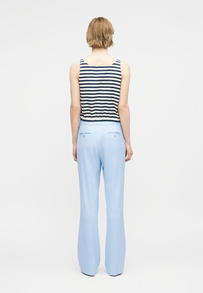 Λεπτός άνθρωπος με κοντά ξανθά μαλλιά, που φοράει αμάνικο μπλε ναυτικό και λευκό ριγέ τοπ και ανοιχτό μπλε παντελόνι με φαρδιά πόδια, γυρισμένος προς τα πίσω.