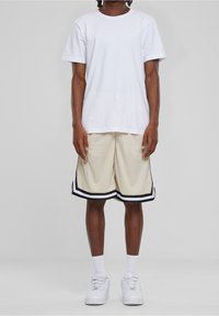 Une personne portant un t-shirt blanc, un short de basketball beige avec des bordures noir et blanc, des chaussettes blanches et des baskets blanches, se tenant sur un sol blanc.