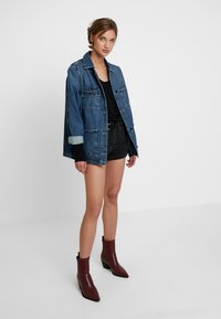 Jaqueta de ganga azul com botões prateados, dois bolsos frontais e punhos em azul claro contrastante; combinada com uma blusa preta e calções pretos. Botas de tornozelo castanhas.