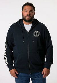 Sweat à zip bleu marine en tissu doux, avec un logo sur le devant, des poches latérales et un texte blanc sur la manche gauche. Design décontracté.