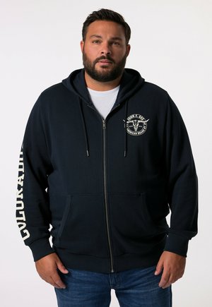 Navy modrá mikina na zip vyrobená z měkké tkaniny, s předním logem, bočními kapsami a bílým textem na levém rukávu. Casual design.