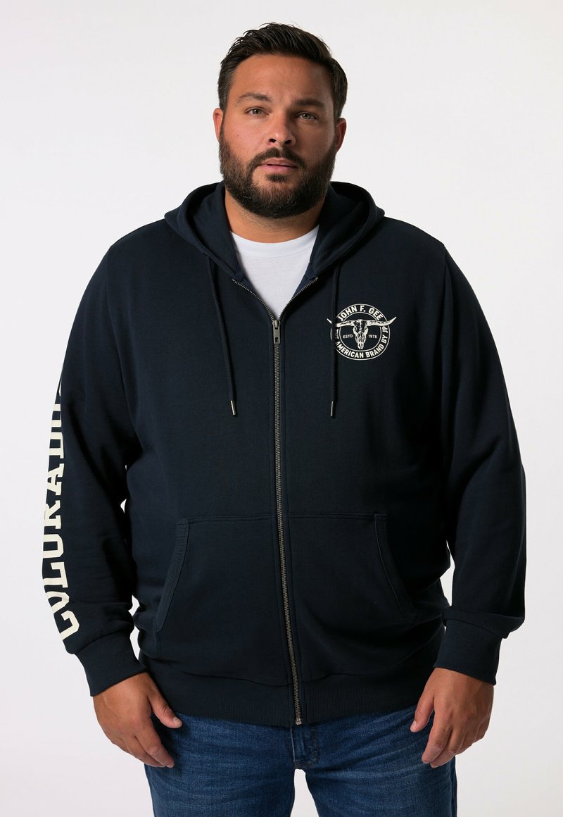 Sweat à zip bleu marine en tissu doux, avec un logo sur le devant, des poches latérales et un texte blanc sur la manche gauche. Design décontracté.