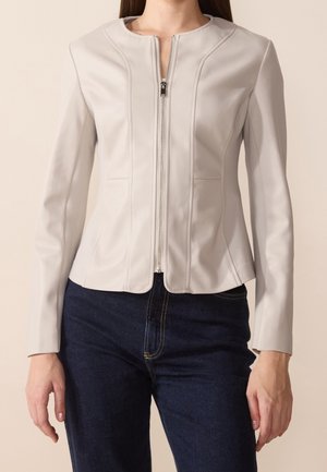 Femme portant une veste légère beige clair ajustée avec fermeture éclair et manches longues, et un jean bleu foncé, debout sur un fond uni.