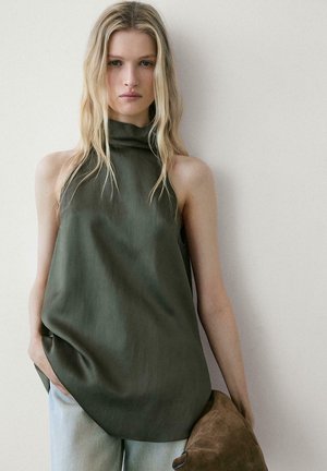 Femme blonde portant un haut sans manches vert foncé à col haut et un jean clair, tenant un chapeau en daim marron contre un fond de mur clair uni.