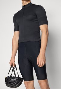 Maillot de cyclisme noir à manches courtes, avec une fermeture éclair à l'avant et une coupe ajustée. Associé à un cuissard rembourré noir et un casque de vélo.