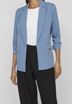 Woman wearing a light blue blazer with three-quarter sleeves, white top, and black high-waisted trousers against a plain background.