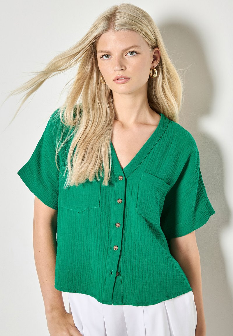 Jeune femme aux longs cheveux blonds portant des boucles d'oreilles dorées en forme de créoles, un chemisier vert à manches courtes boutonné et un pantalon blanc, debout devant un mur clair.