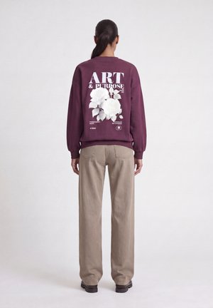 Persoon die een bordeauxrode trui draagt met witte bloemenprint en de tekst "ART & PURPOSE" op de achterkant, beige broek en zwarte schoenen.