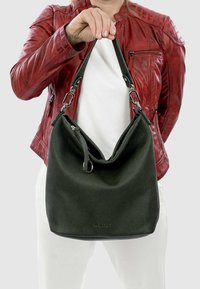 Persona con giacca di pelle rossa e pantaloni bianchi che tiene una borsa di pelle nera per il manico, su uno sfondo semplice.