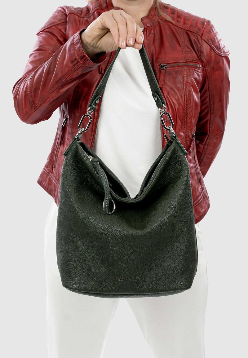 Persona con giacca di pelle rossa e pantaloni bianchi che tiene una borsa di pelle nera per il manico, su uno sfondo semplice.