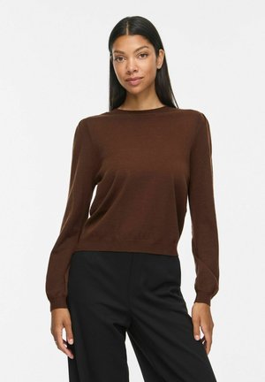 Femme aux longs cheveux noirs porte un pull marron à manches longues et un pantalon noir, debout devant un fond blanc uni.