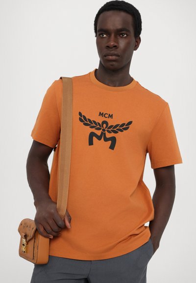 Oranž puuvillane T-särk musta MCM logo graafikuga, ümar kaelus, lühikesed varrukad, koos pruuni mustrilise õlakotiga.