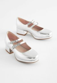 Silberne, metallische Mary Jane Schuhe mit zwei Riemen und runder Zehenpartie, die einen klobigen Absatz und eine glatte Oberflächenstruktur aufweisen.