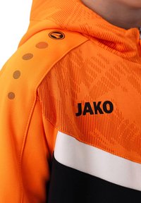Oranger Hoodie aus strukturiertem Material, mit einem schwarzen Logo "JAKO" und weißen Akzenten. Enthält dekorative kreisförmige Muster auf der Schulter.