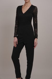 Combinaison noire avec un décolleté en V, des manches longues en dentelle et une taille cintrée. Les jambes sont fuselées et fabriquées dans un tissu lisse. Pas de motifs ni de quincaillerie.