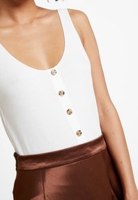 Bílý žebrovaný top s hlubokým výstřihem a čtyřmi zlatými knoflíky v centru, doplněný lesklou hnědou saténovou sukní.